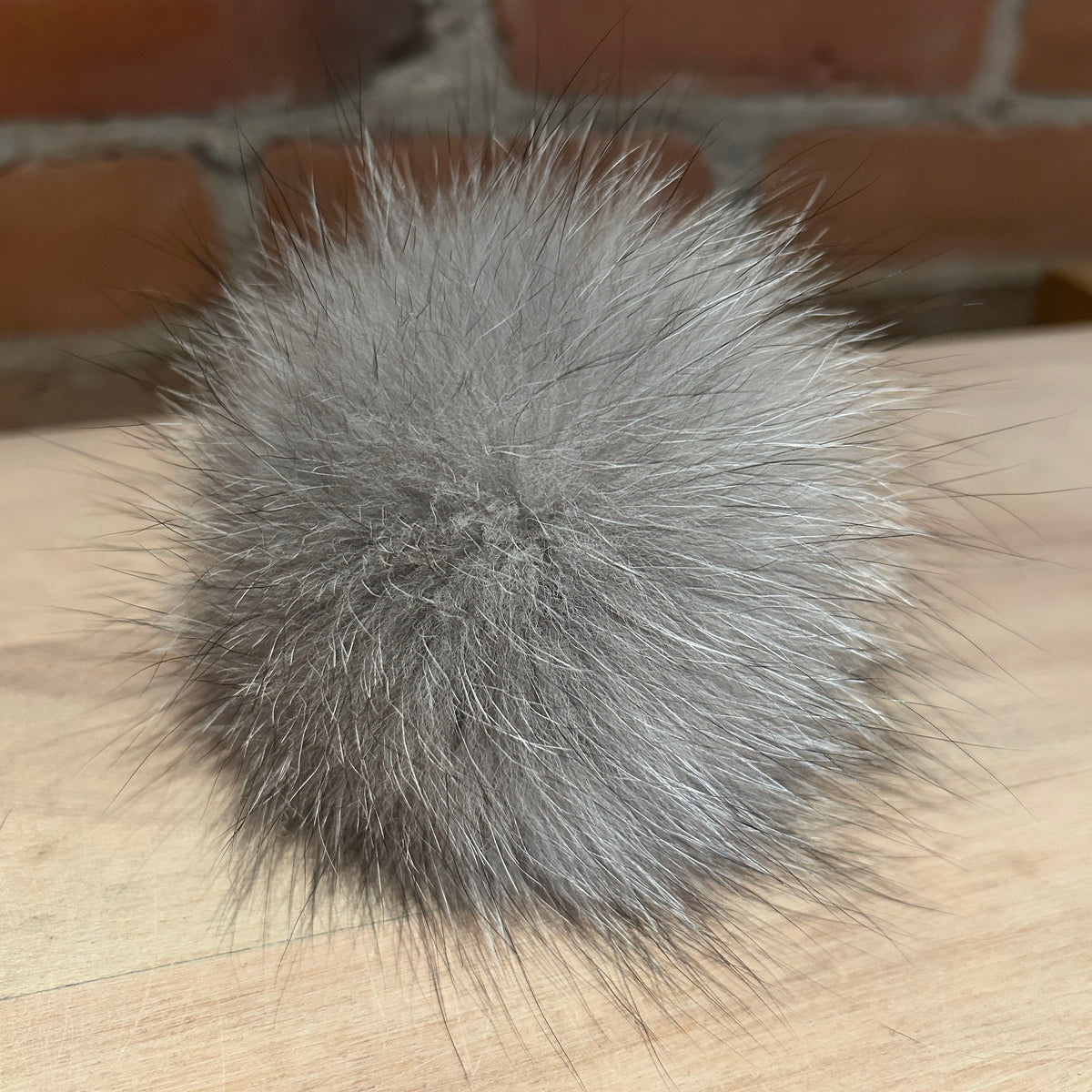A fluffy gray fox fur pom-pom ball resting on a light wood surface with a brick wall background.