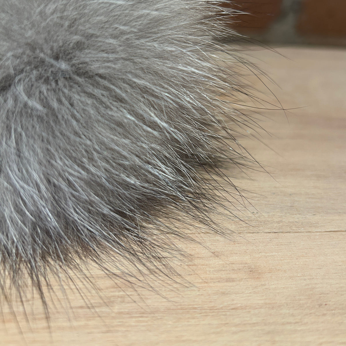 A close-up of a fluffy gray fox fur pom-pom resting on a light wood surface with a blurred brick wall in the background.