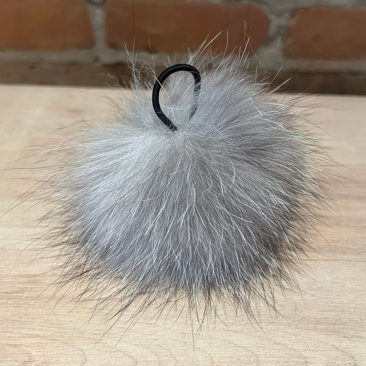 A fluffy gray fox fur pom-pom keychain or bag charm with a black plastic loop at the top, resting on a light wood surface with a brick wall in the background.