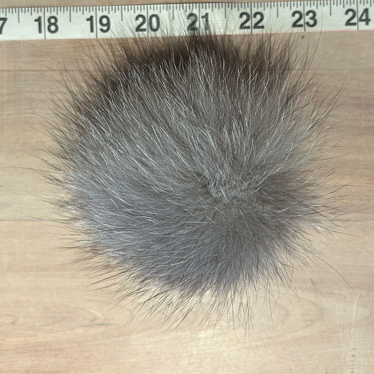 A round gray fox fur pom-pom with dark tips, shown on a light wood surface next to a measuring tape ruler for scale.