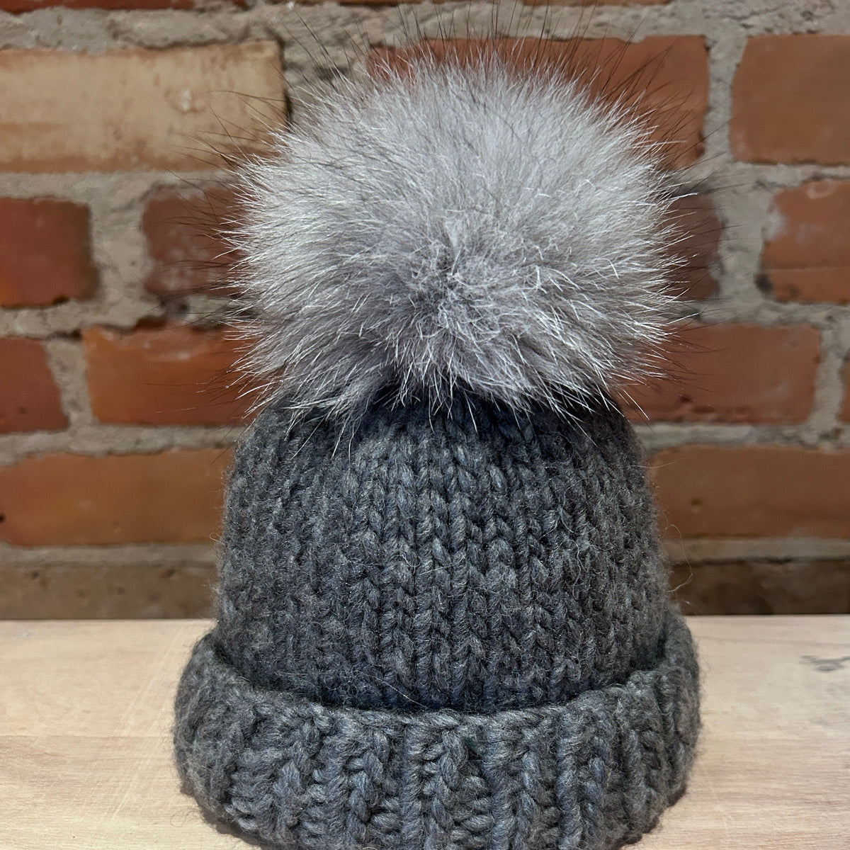 A dark gray knit beanie hat with a large fluffy light gray pom-pom, displayed on a light wood surface against a brick wall background.