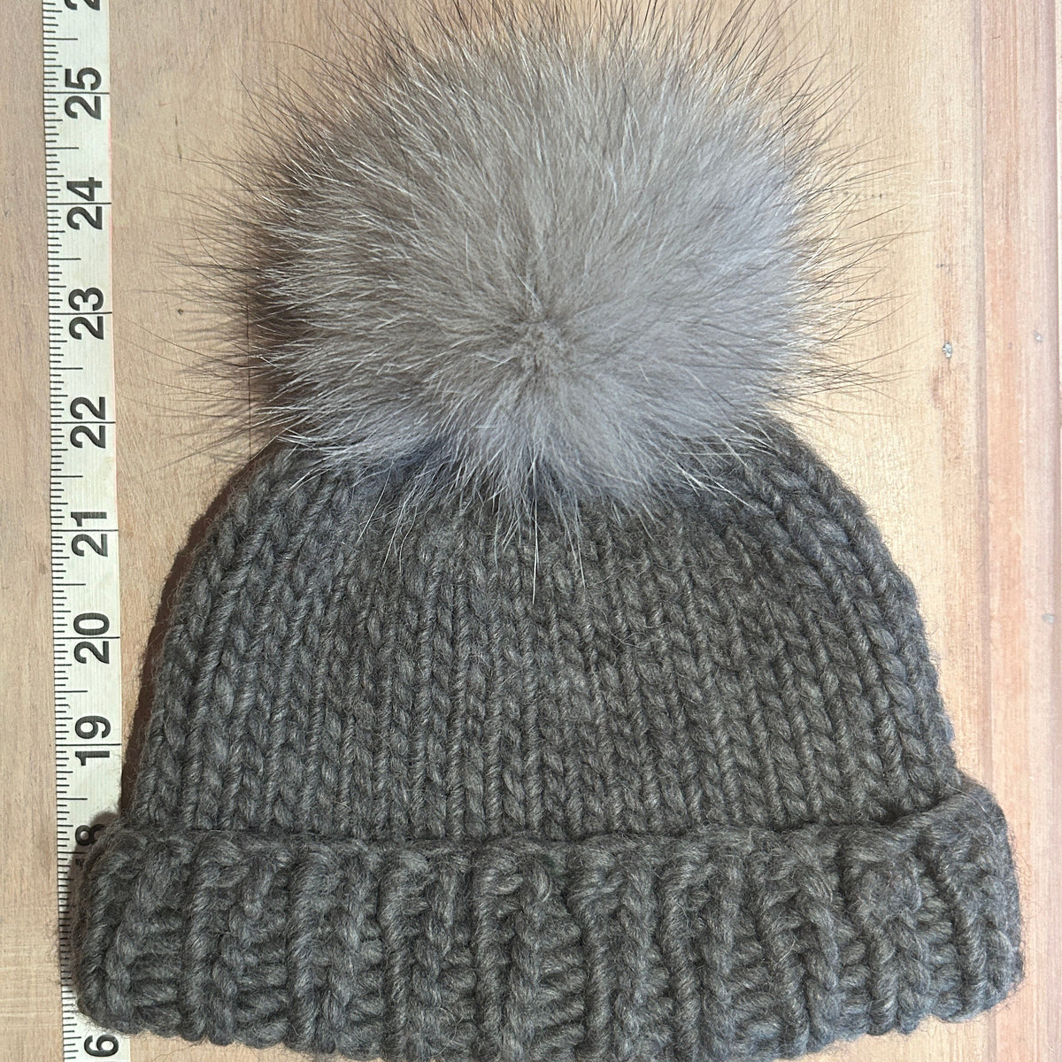 A small gray knit beanie with a small fluffy gray fox-fur pom-pom, laid flat on a light wood surface next to a measuring ruler.