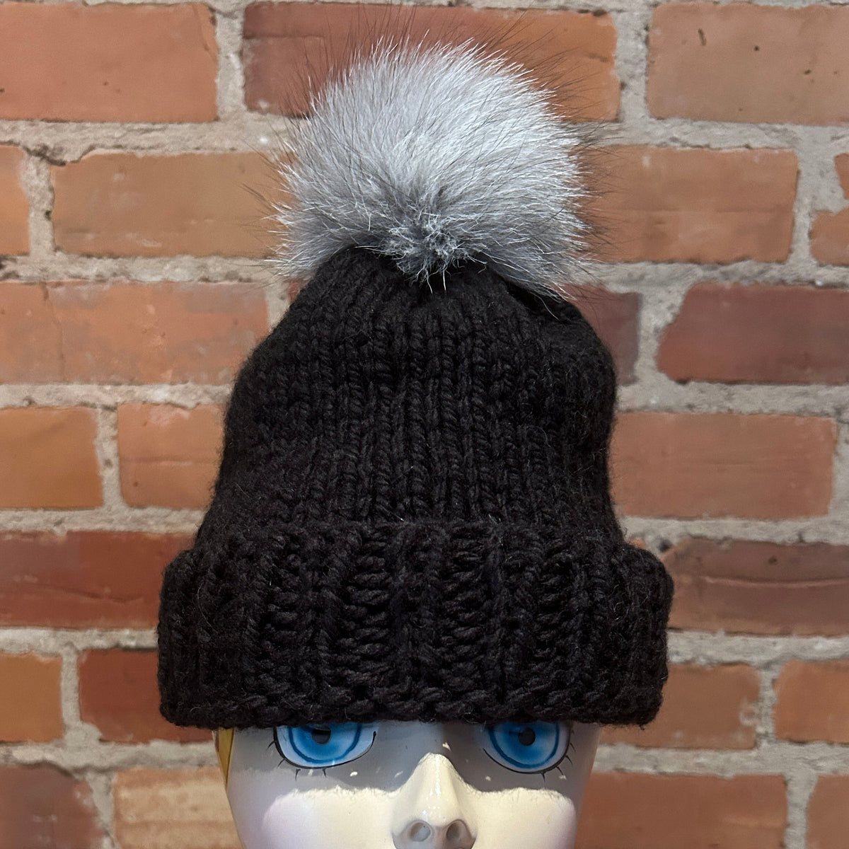 A black ribbed knit beanie hat with a fluffy light gray pom-pom, displayed on a mannequin head against a red brick wall background.