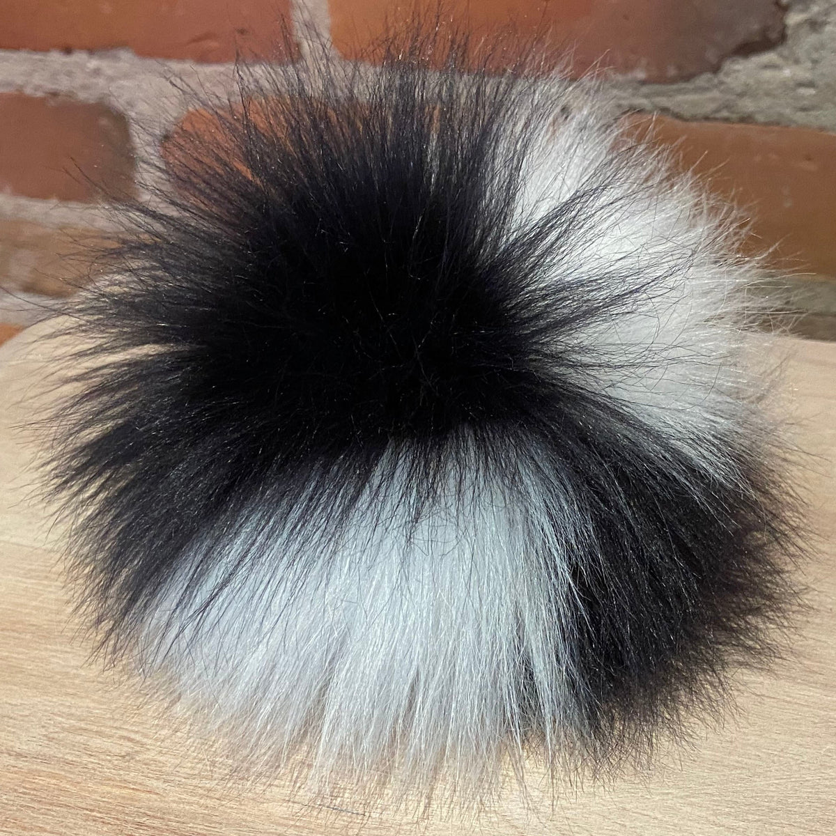A fluffy faux-fur pom-pom accessory with a black and white color-blocked design, resting on a light wood surface with a brick wall background.