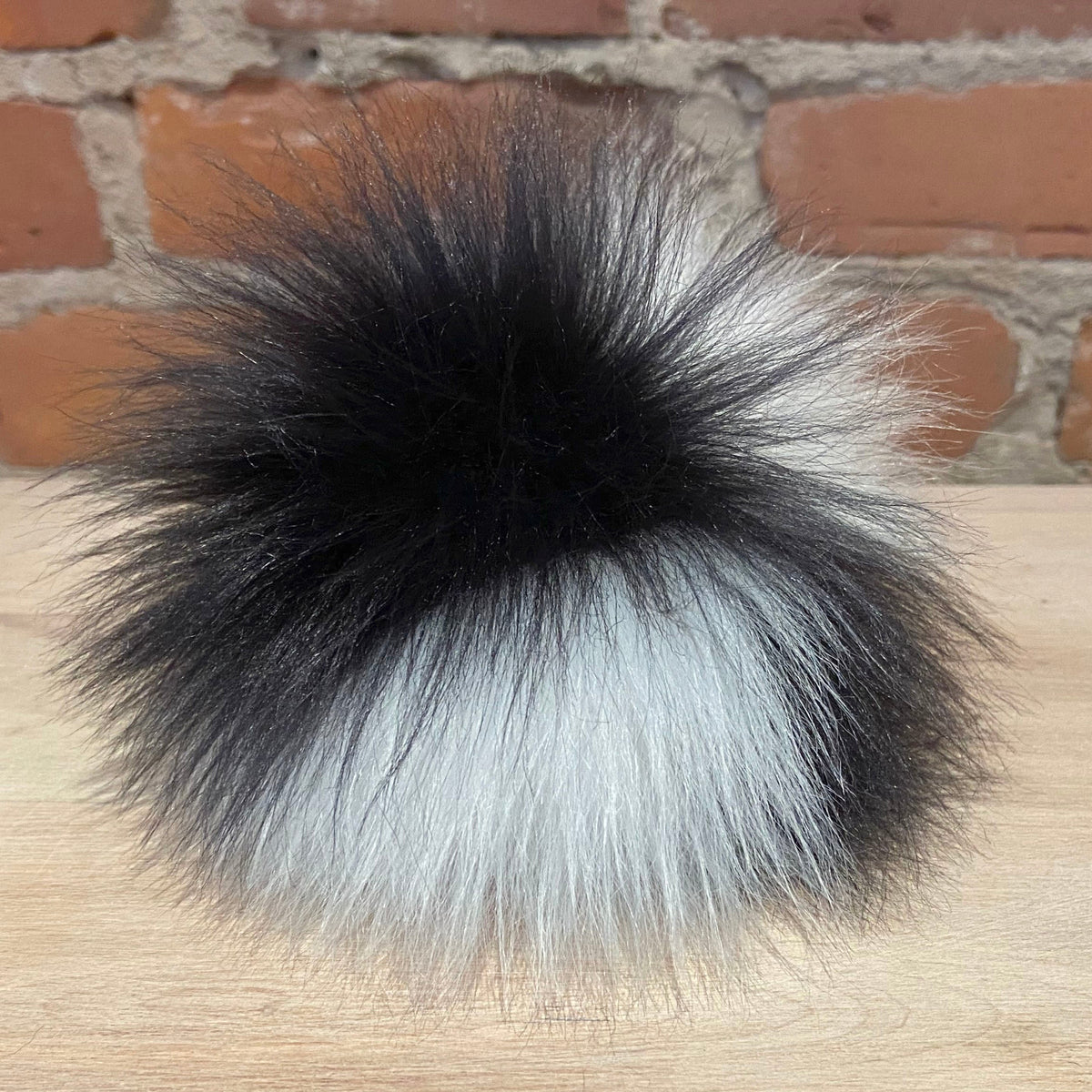 A fluffy faux fur pom-pom accessory with a black top section and a white bottom section, resting on a light wood surface with a brick wall background.