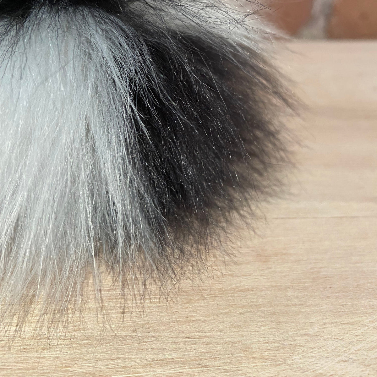 A close-up of a fluffy faux fur pom-pom with black and silver-gray fibers resting on a light wood surface.