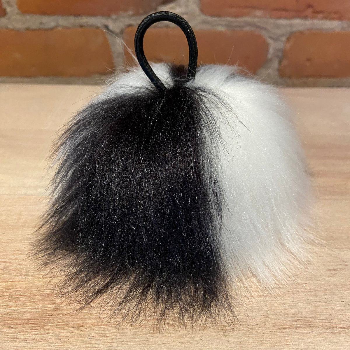 A fluffy faux fur keychain or bag charm with a black and white split design, featuring a black elastic loop at the top, resting on a light wood surface with a brick wall background.