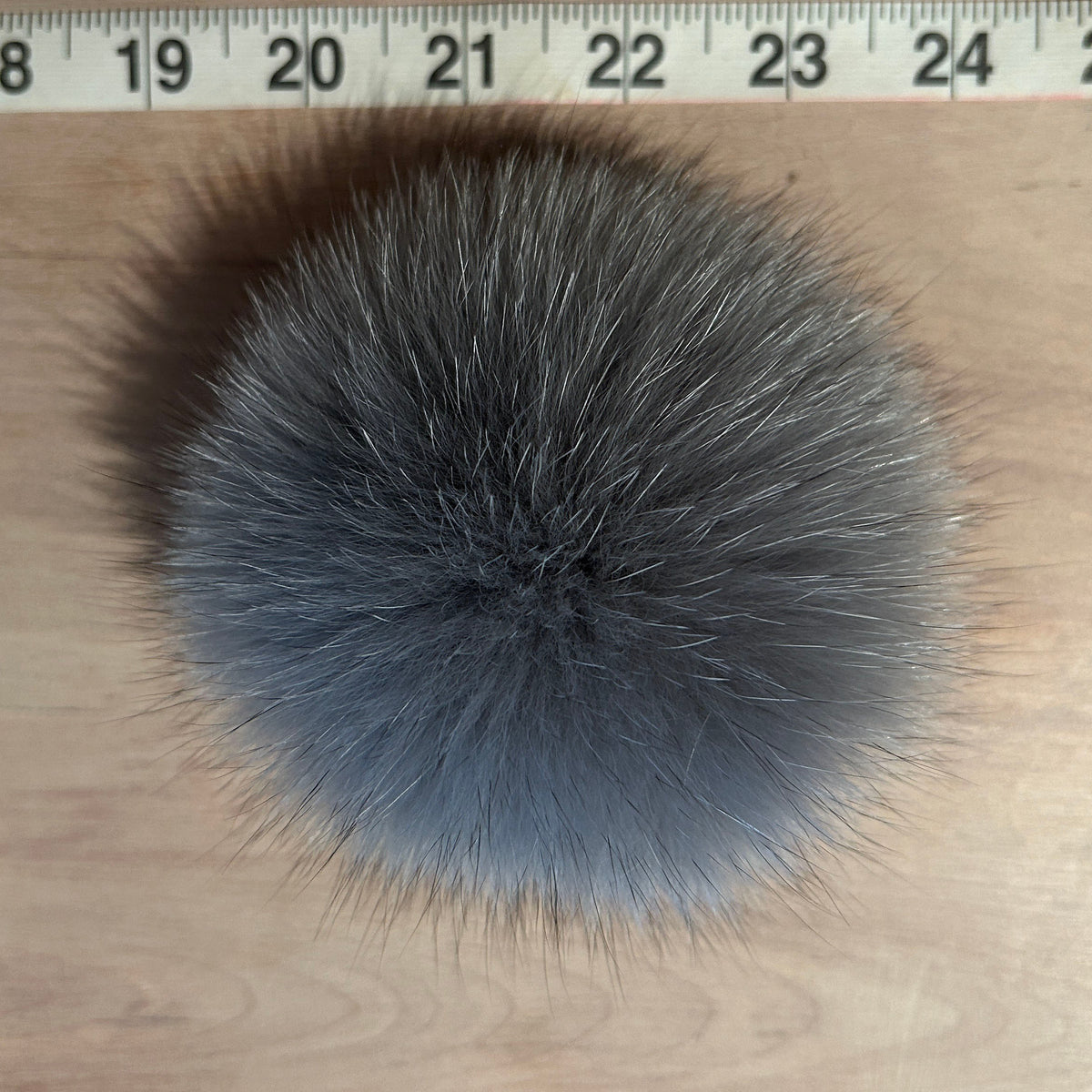 A round gray fox fur pom-pom with black and white mixed fur tips, shown on a wooden surface with a measuring tape at the top for scale.