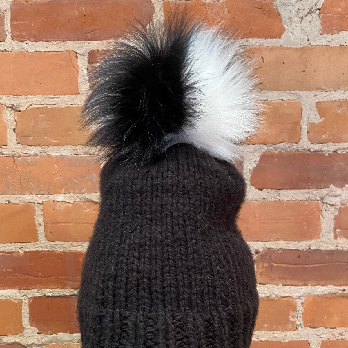 A close-up back view of a black ribbed knit beanie with a large fluffy pom-pom in black and white, set against a red brick wall background.