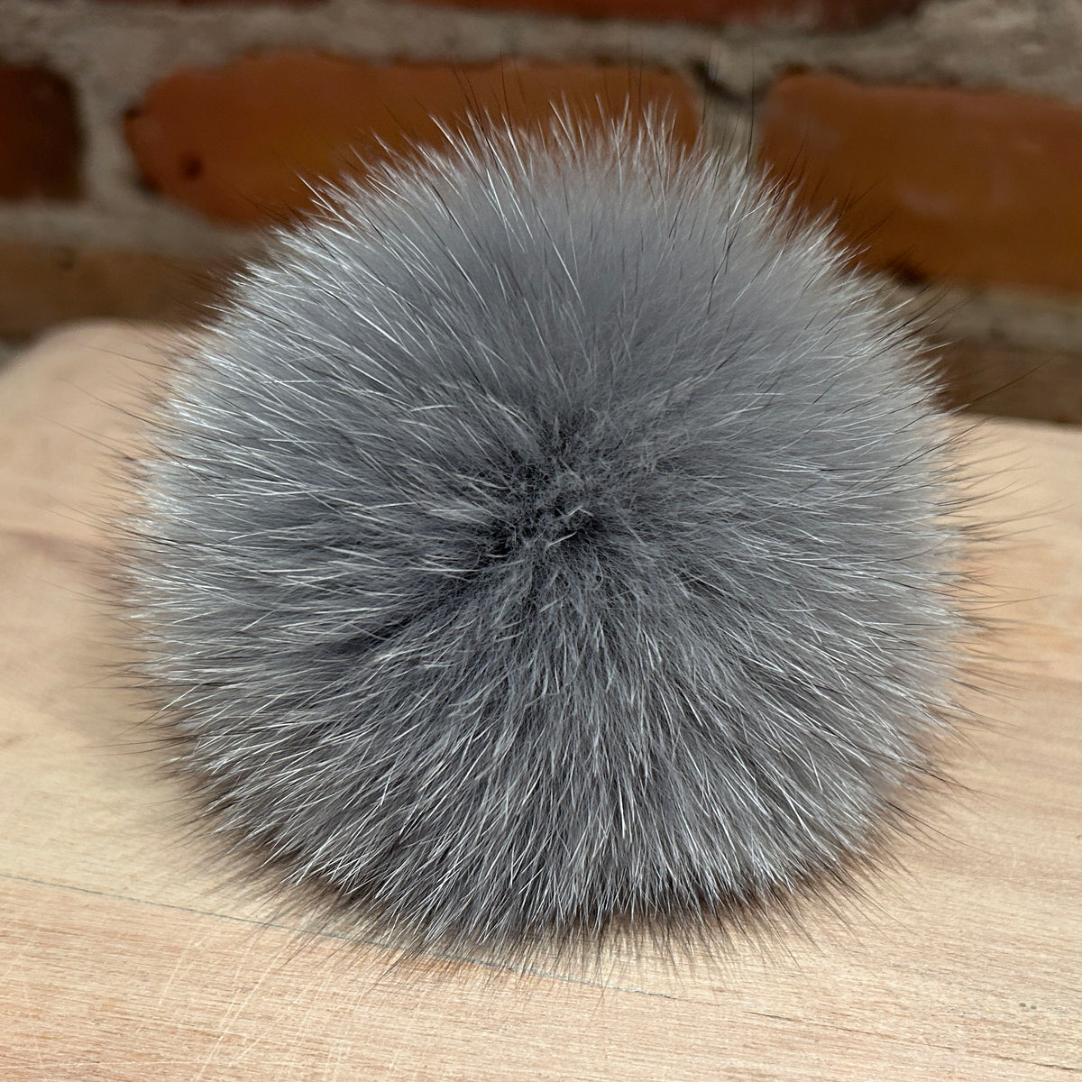 A fluffy gray fox fur pom-pom ball resting on a light wood surface with a blurred brick wall background.