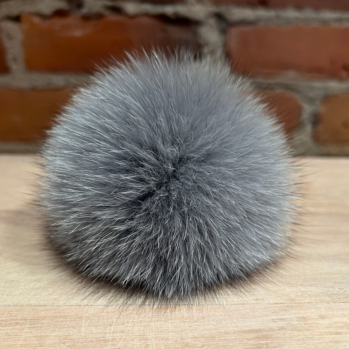 A round, fluffy gray fox fur pom-pom accessory resting on a light wood surface with a blurred brick wall background.