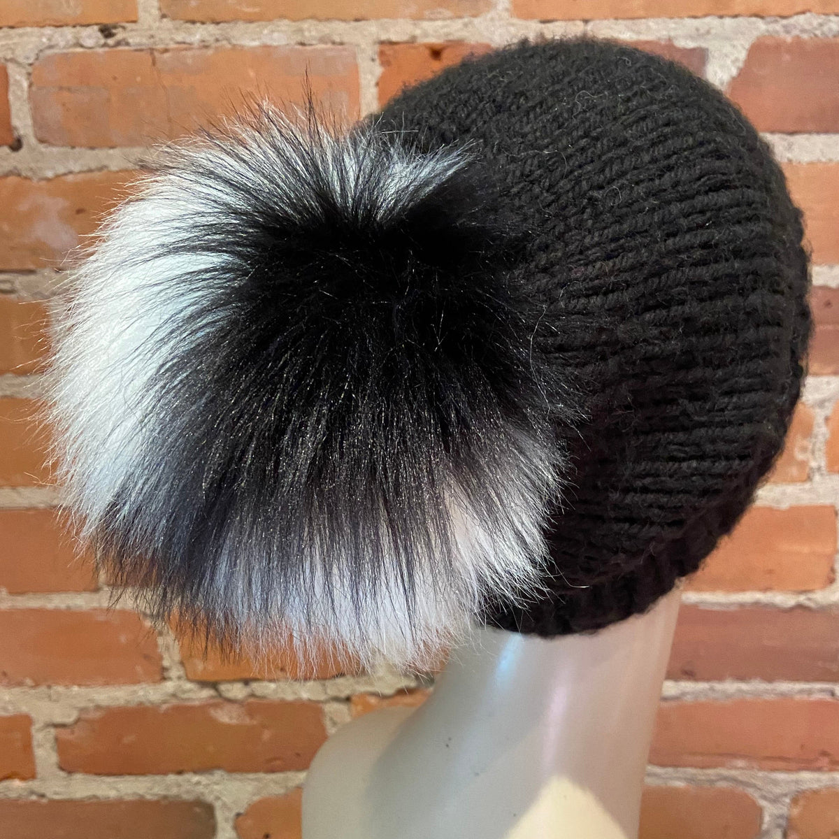 A black ribbed knit beanie hat with a large fluffy pom-pom in black, white, and gray, displayed on a mannequin head against a brick wall background.