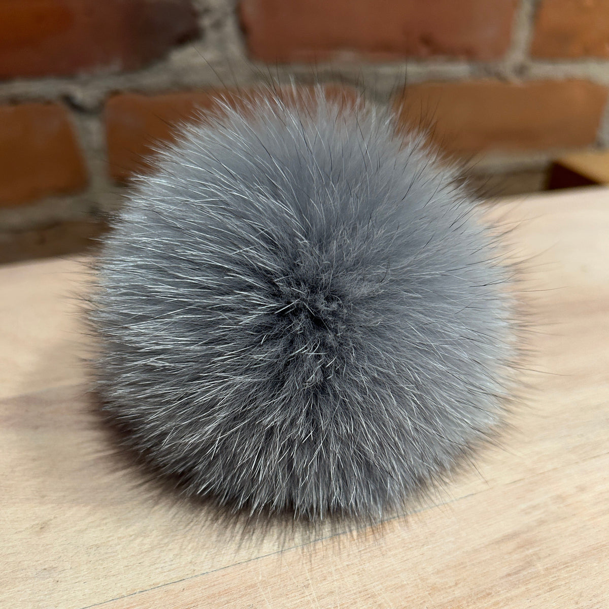 A fluffy gray fox fur pom-pom ball resting on a light wood tabletop with a blurred brick wall background.