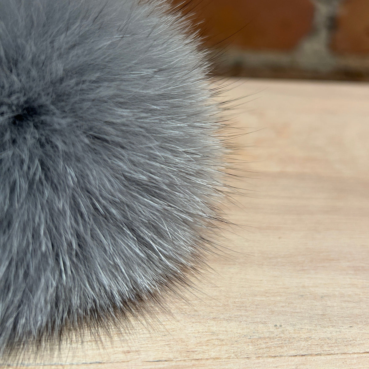 A close-up of a fluffy gray fox fur pom-pom resting on a light wood surface with a blurred red brick wall background.