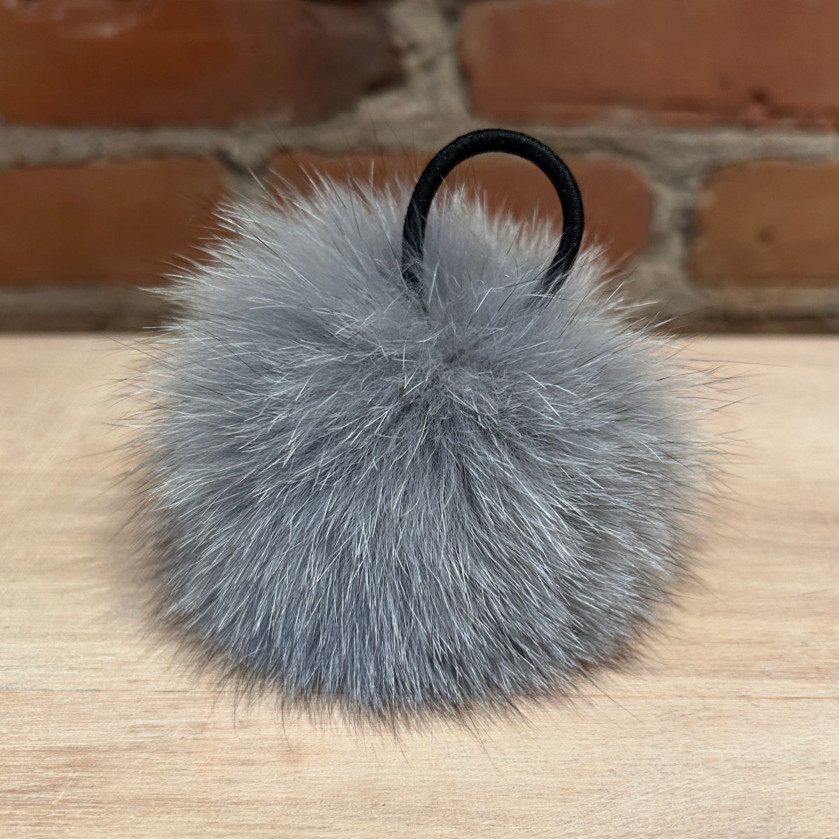 A gray fox fur pom-pom keychain or bag charm with a black elastic loop, resting on a light wood surface with a brick wall background.