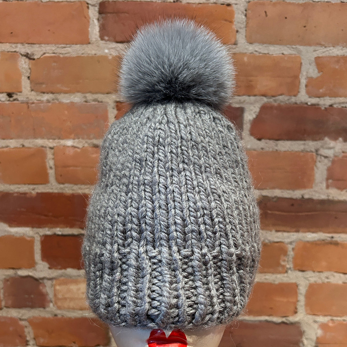 A gray chunky knit beanie with a matching gray fox fur pom-pom, displayed on a mannequin head against a red brick wall background.