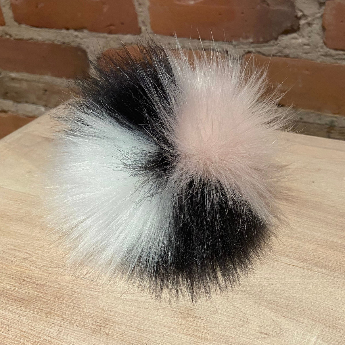 A fluffy pom-pom hair accessory with a color-blocked design in black, white, and pale pink, resting on a light wood surface with a brick wall background.