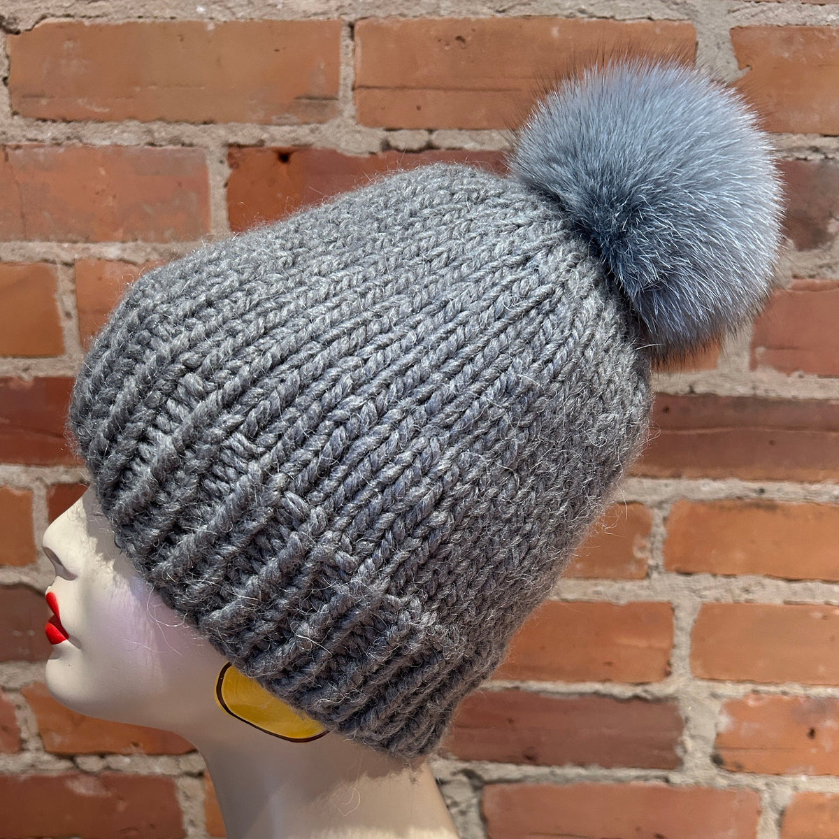 A gray ribbed knit beanie with a fluffy pom-pom, displayed on a mannequin head against a red brick wall background.