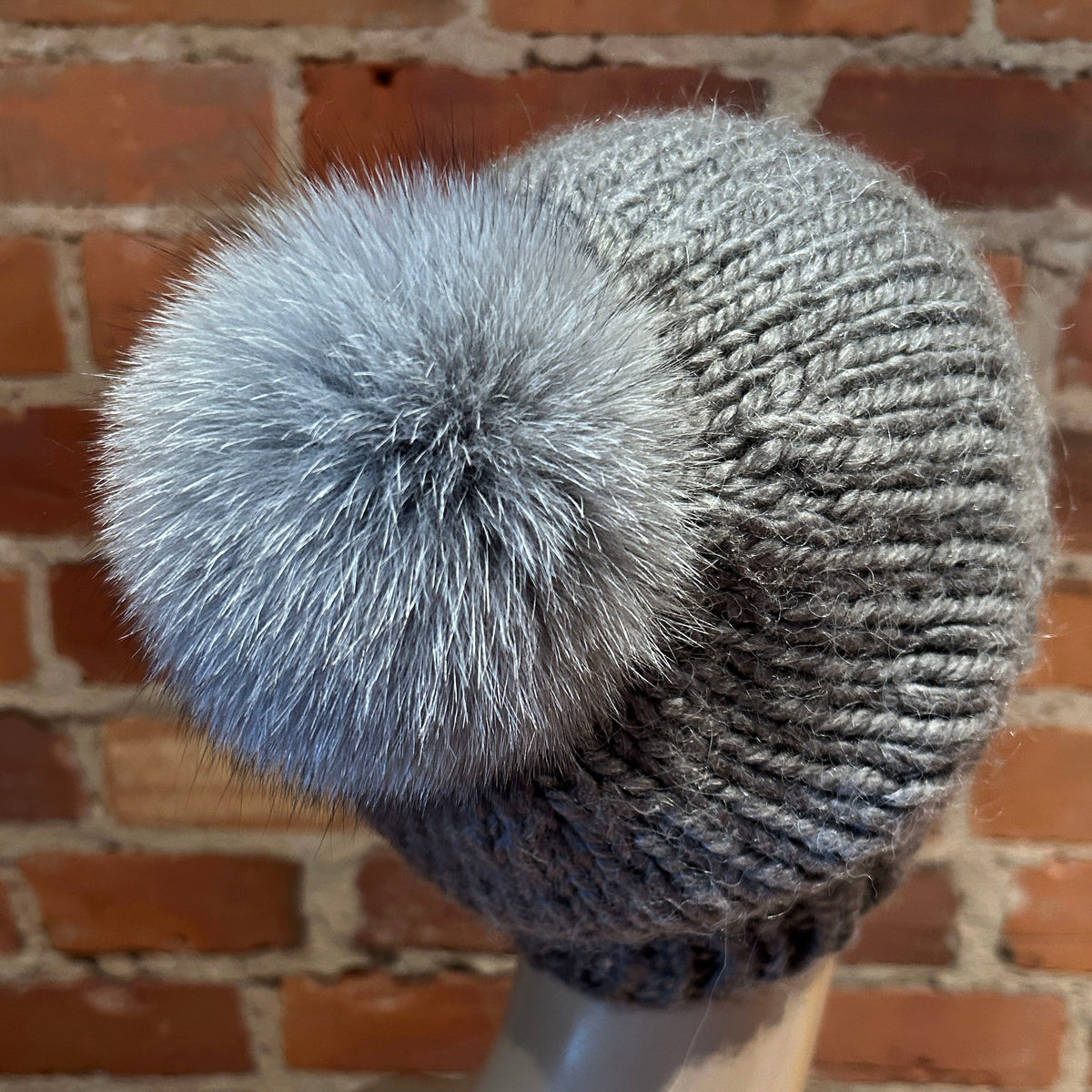 A gray knit beanie with a large fluffy gray fox-fur pom-pom, displayed on a mannequin head against a red brick wall background.