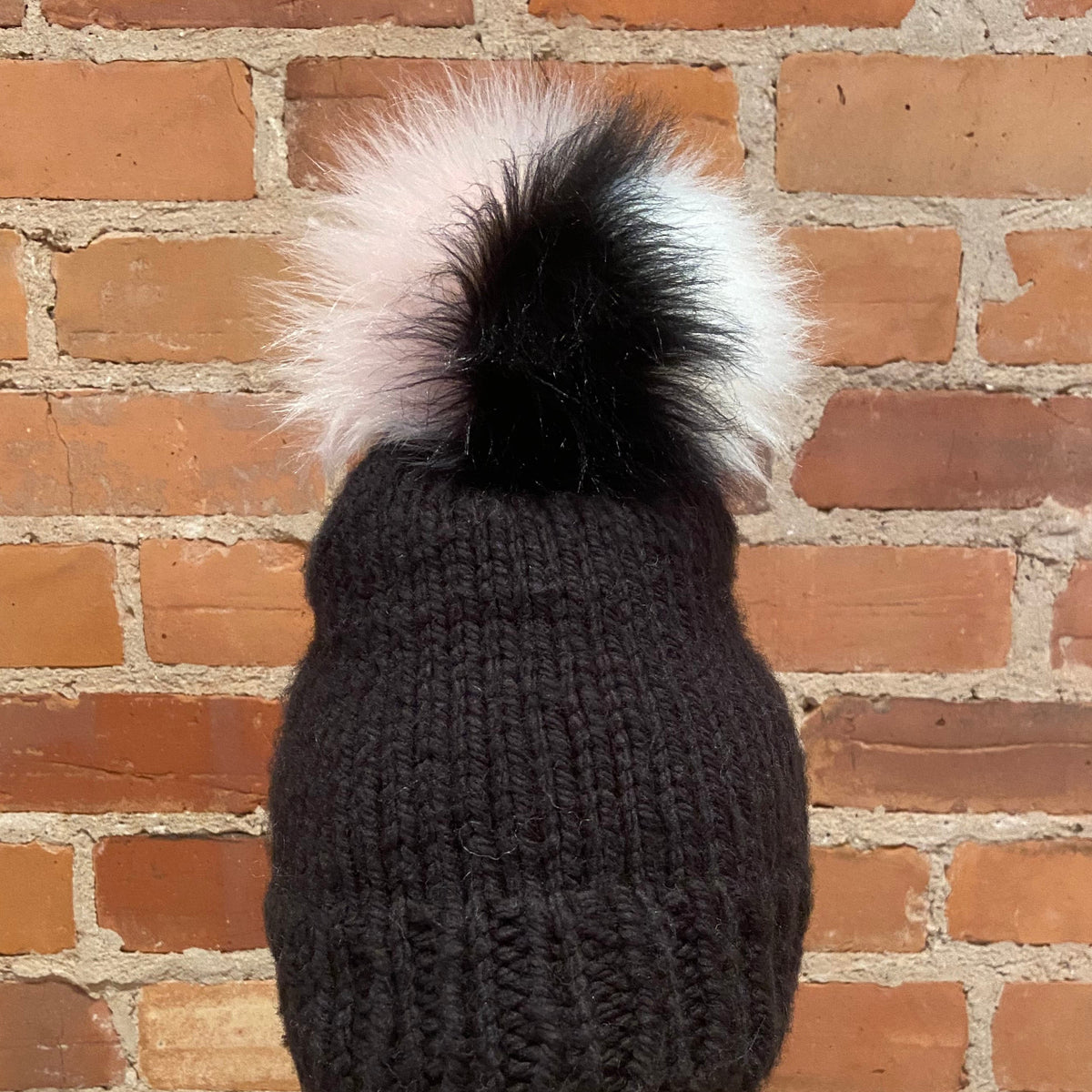 A close-up of a dark charcoal knit beanie with a large fluffy pom-pom in black, white, and light pink, set against a red brick wall background.
