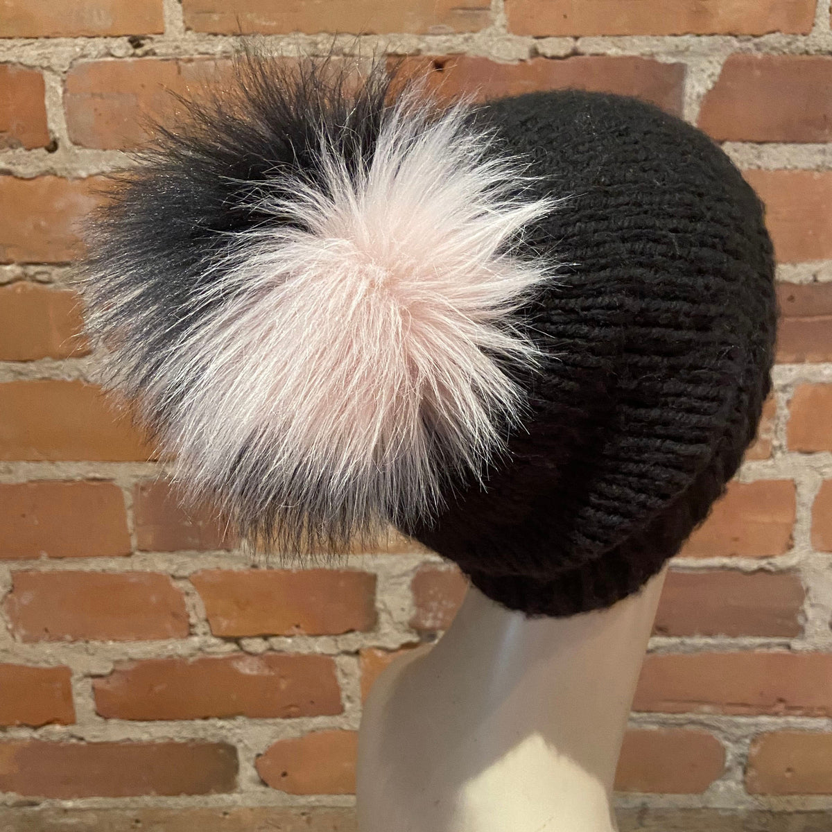 A black ribbed knit beanie hat with a large fluffy pom-pom in black, white, and pale pink, displayed on a mannequin head against a brick wall background.