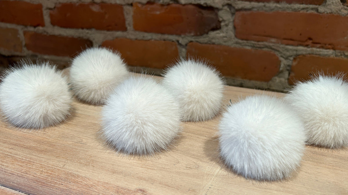 Six fluffy white faux-fur pom-poms arranged on a light wood surface in front of a red brick wall.