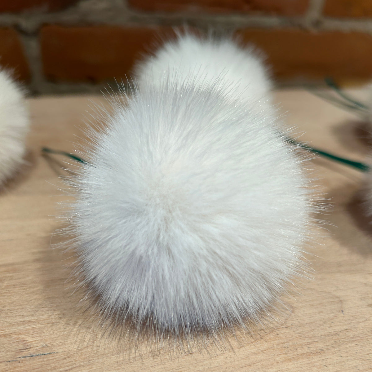 A close-up of a fluffy white faux fur pom-pom christmas tree ornament on a light wood surface with a brick wall background.