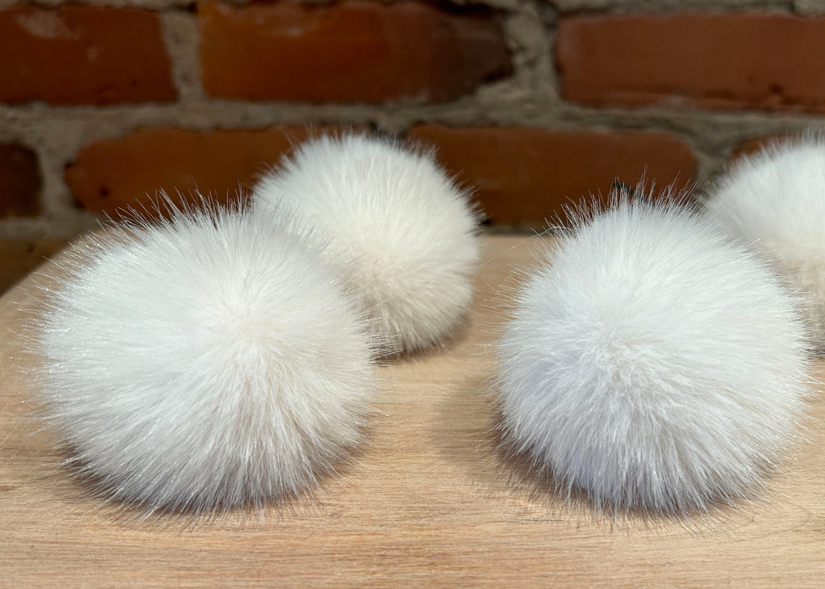 Four fluffy white faux-fur pom-poms arranged on a light wood surface with a brick wall background.