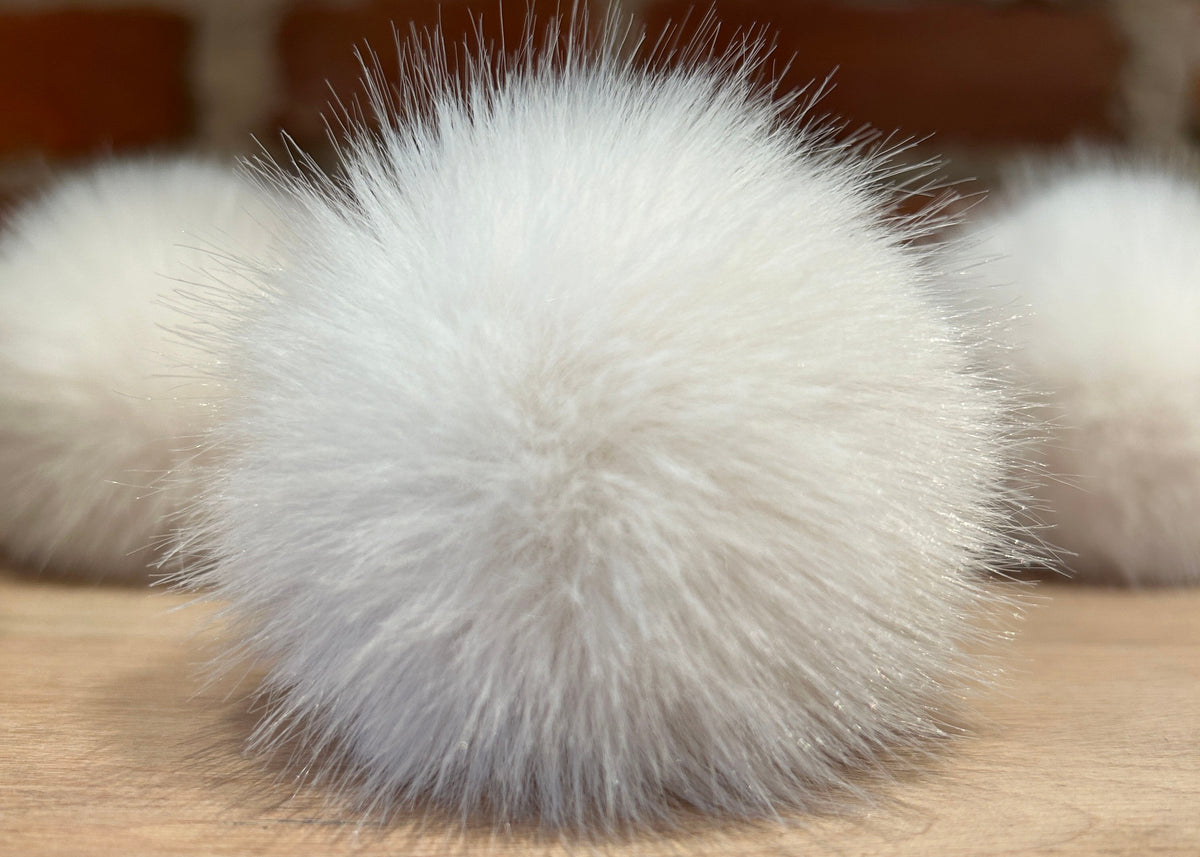 A fluffy white faux fur pom-pom ball resting on a light wood surface with a blurred brick wall background.