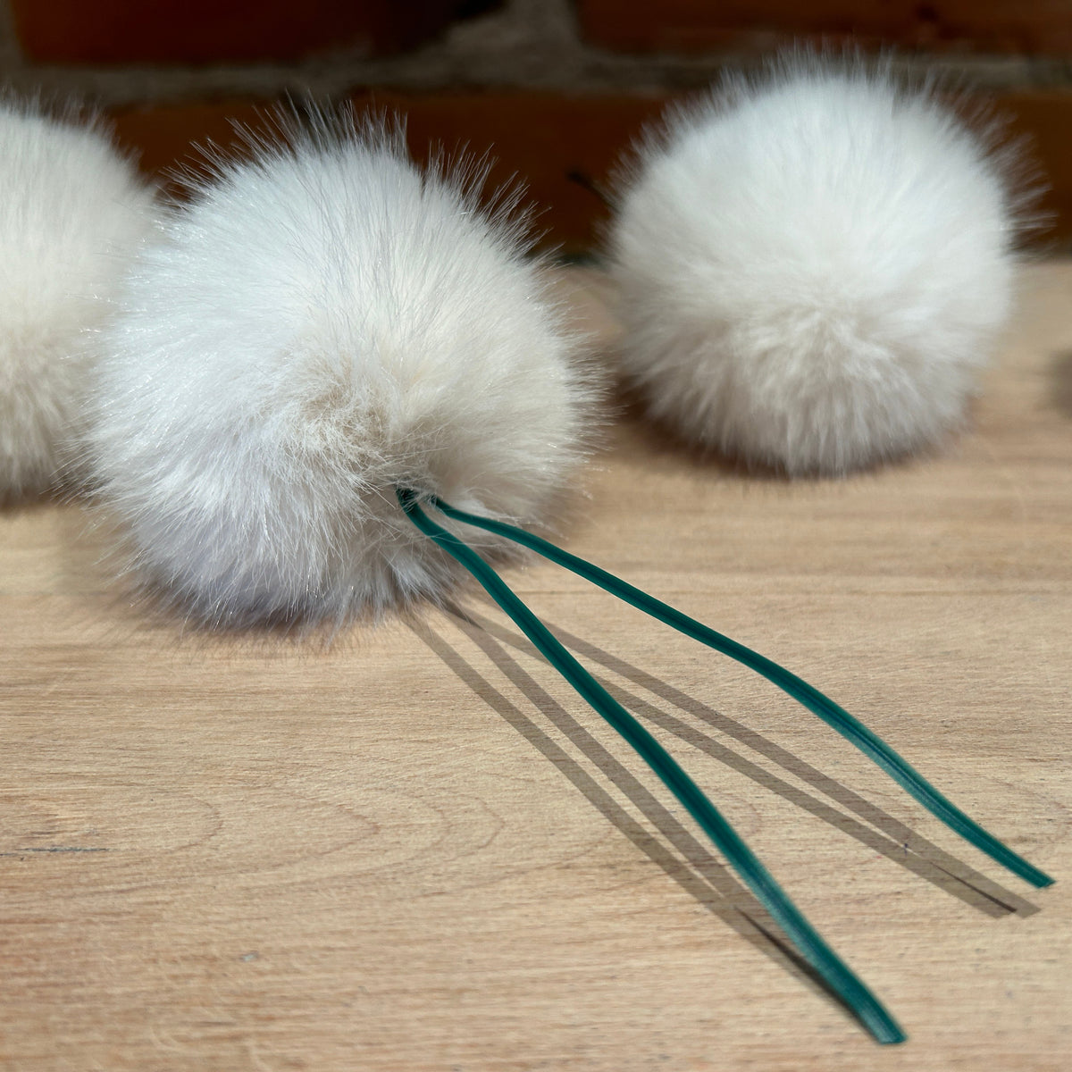 Three fluffy white faux fur pom-poms with long dark green twist tie tails, arranged on a light wood surface.