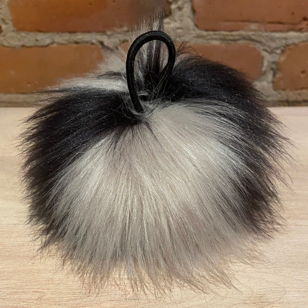 A fluffy faux fur pom-pom keychain or bag charm in black and silver-gray, attached to a black elastic loop, resting on a light wood surface with a brick wall background.