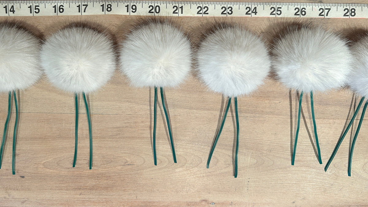 Six white fluffy pom-pom christmas tree ornaments with dark green wire stems arranged in a row on a light wood surface, with a measuring tape visible at the top for scale.