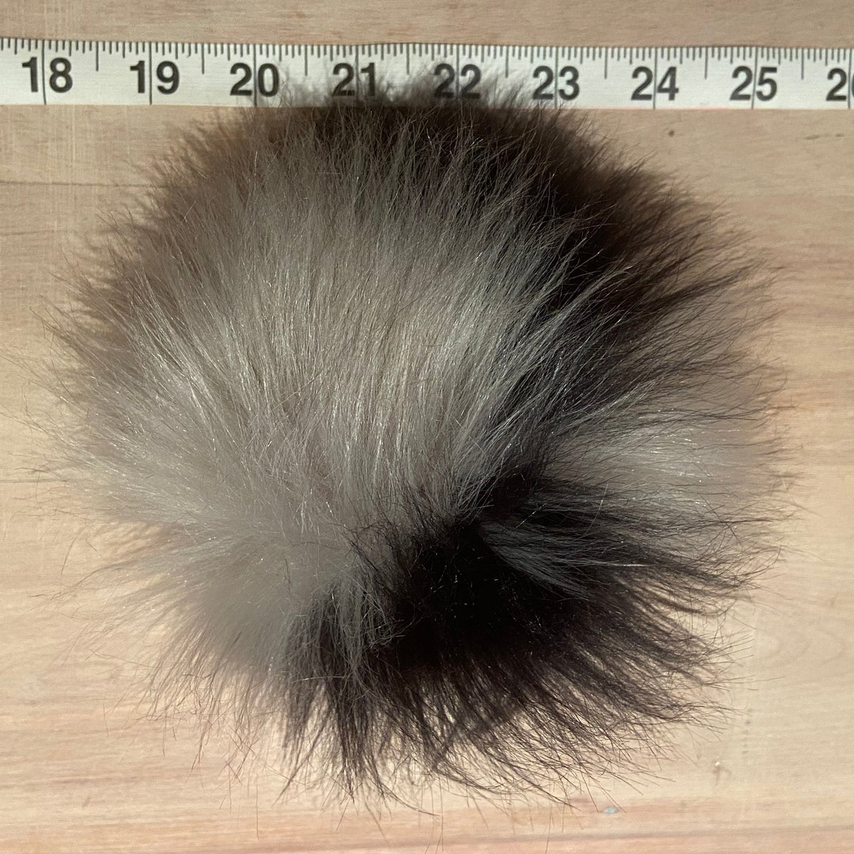 A fluffy pom-pom ball made of black and gray faux fur, shown on a light wood surface with a measuring tape ruler at the top for scale.