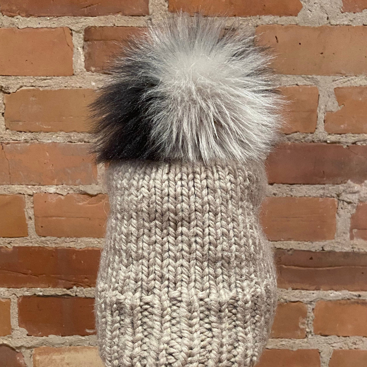 A chunky knit beanie hat in a light oatmeal beige color, featuring a large fluffy pom-pom on top with a black-to-white gradient transition. The hat is displayed on a mannequin head with red lips, set against a rustic red-brown brick wall background.
