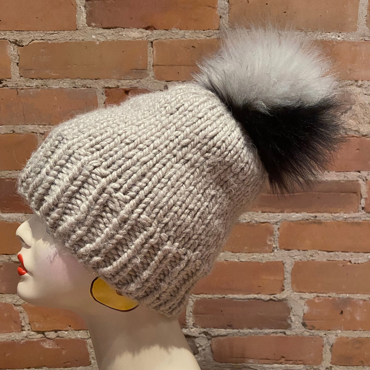 A chunky knit beanie hat in a light oatmeal beige color, featuring a large fluffy pom-pom with a black base and white tip, displayed on a mannequin head against a brick wall background.