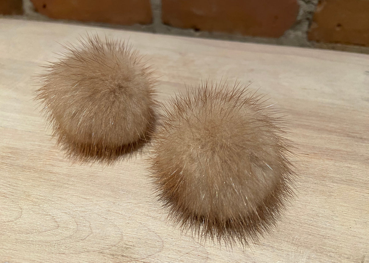 Two round, fluffy mink-fur pom-poms in a light brown/tan color, resting on a light wood surface with a brick wall in the background.