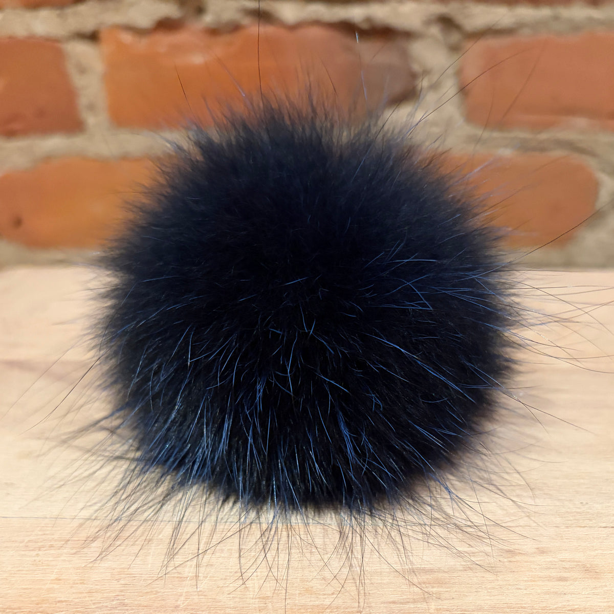 A fluffy indigo blue fox fur pom-pom with subtle blue undertones, resting on a light wood surface with a blurred brick wall background.