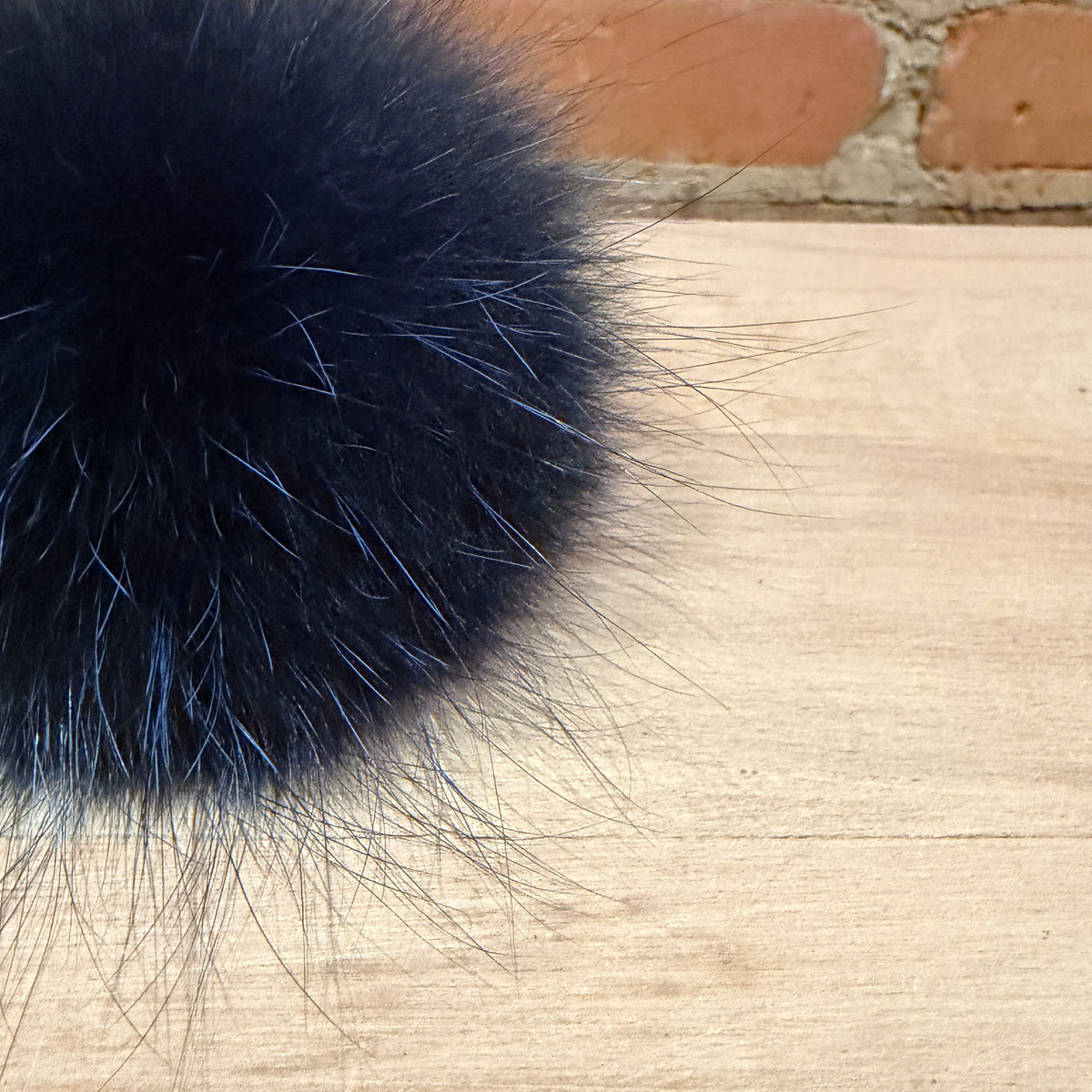 A close-up of a fluffy indigo blue fox fur pom-pom resting on a light wood surface with a brick wall in the background.