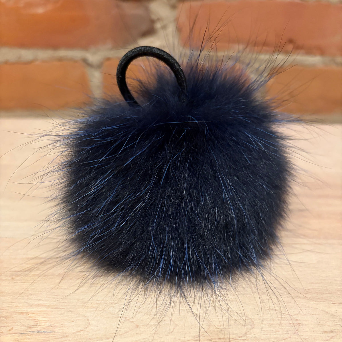 A dark indigo blue fox fur pom-pom keychain or bag charm with a black elastic loop, resting on a light wood surface with a brick wall background.