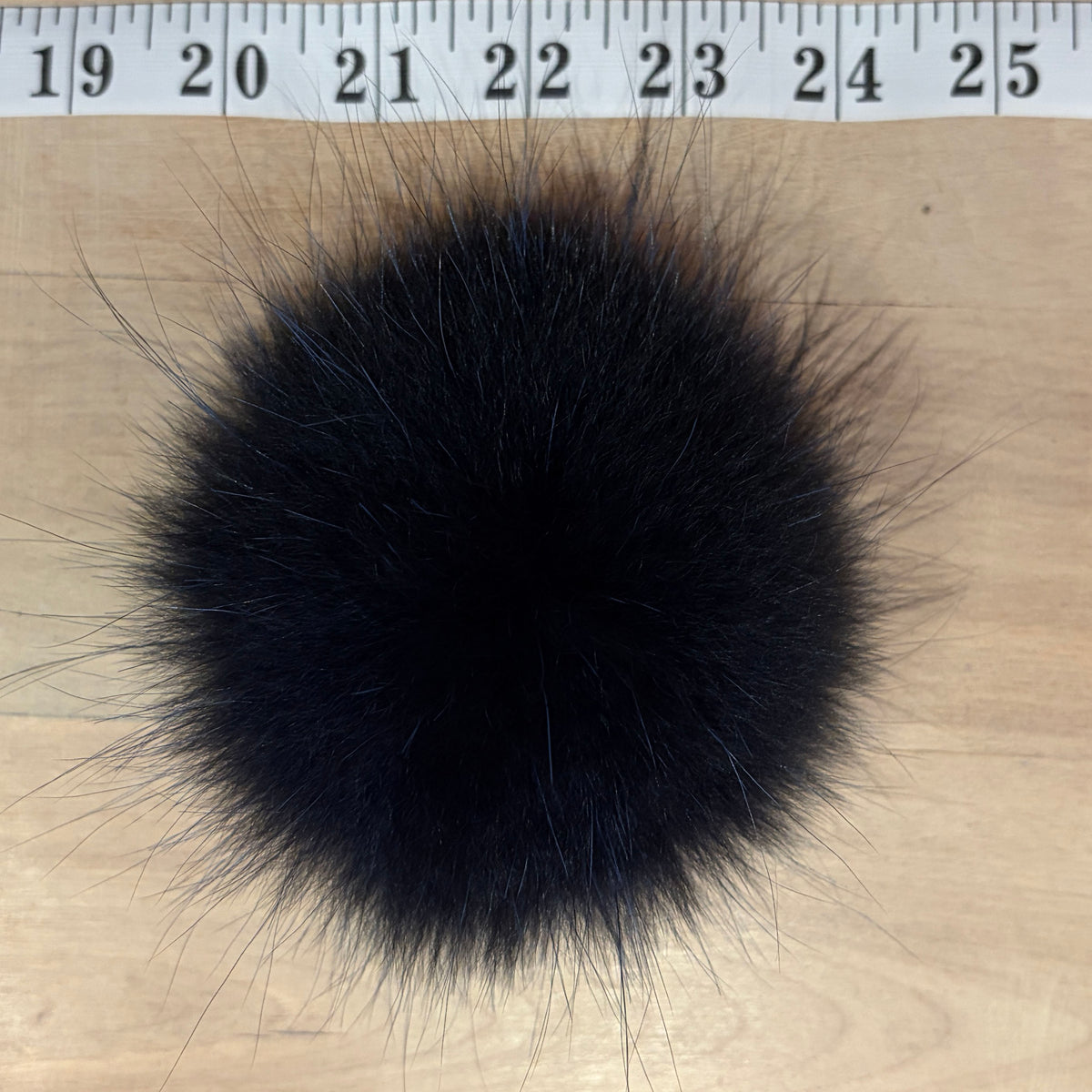 A round indigo blue fox fur pom-pom with wispy, spiky strands, shown on a light wood surface with a measuring tape ruler at the top for scale.