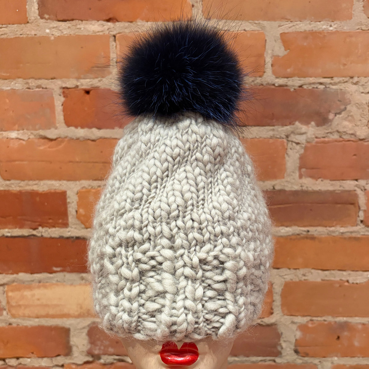 A chunky knit beanie hat in a light oatmeal gray color, topped with a indigo blue fox fur pom-pom. The hat is displayed on a mannequin head with red lips, set against a rustic red brick wall background.