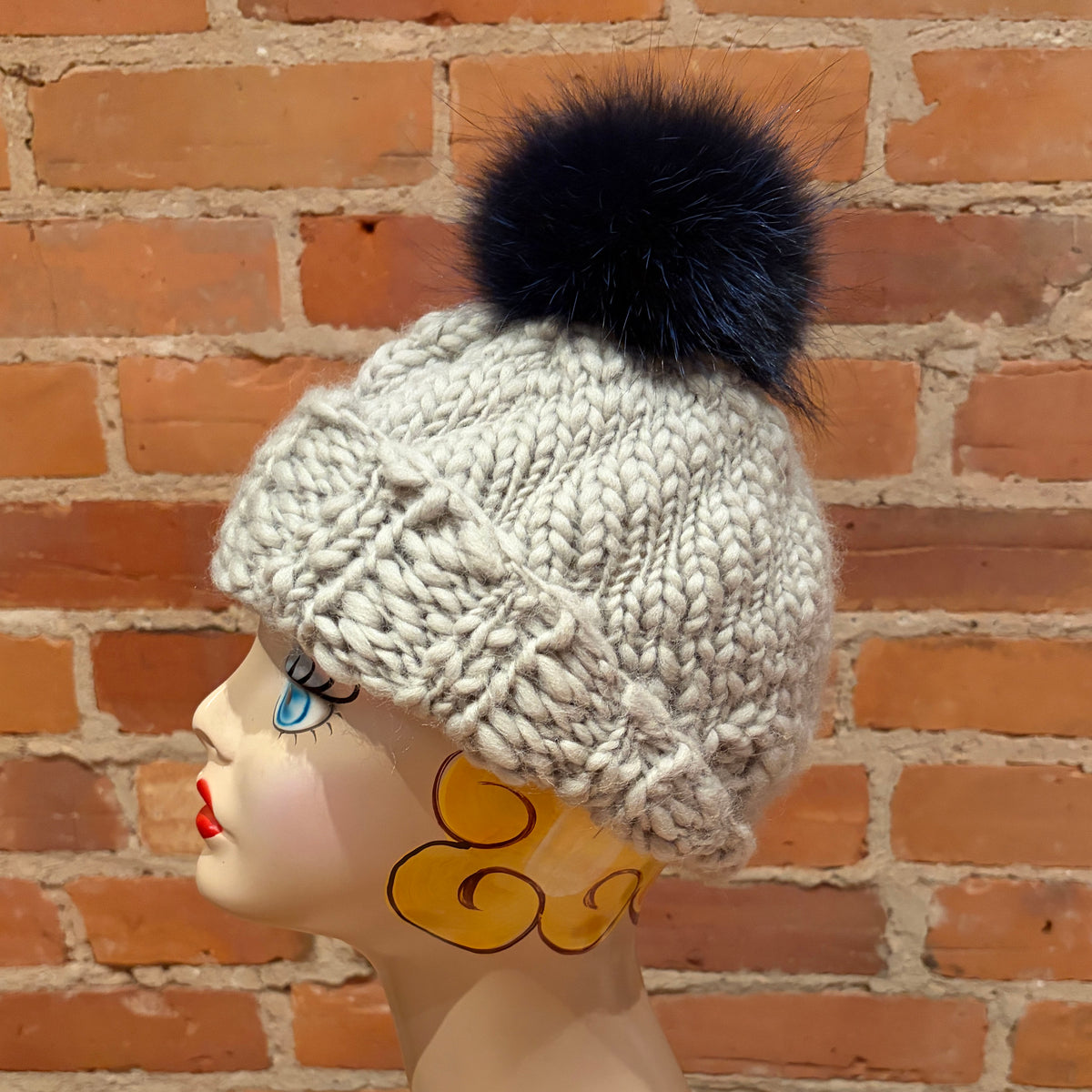 A chunky knit winter beanie in light gray with a indigo blue fox fur pom-pom, displayed on a decorative mannequin head against a red brick wall background.