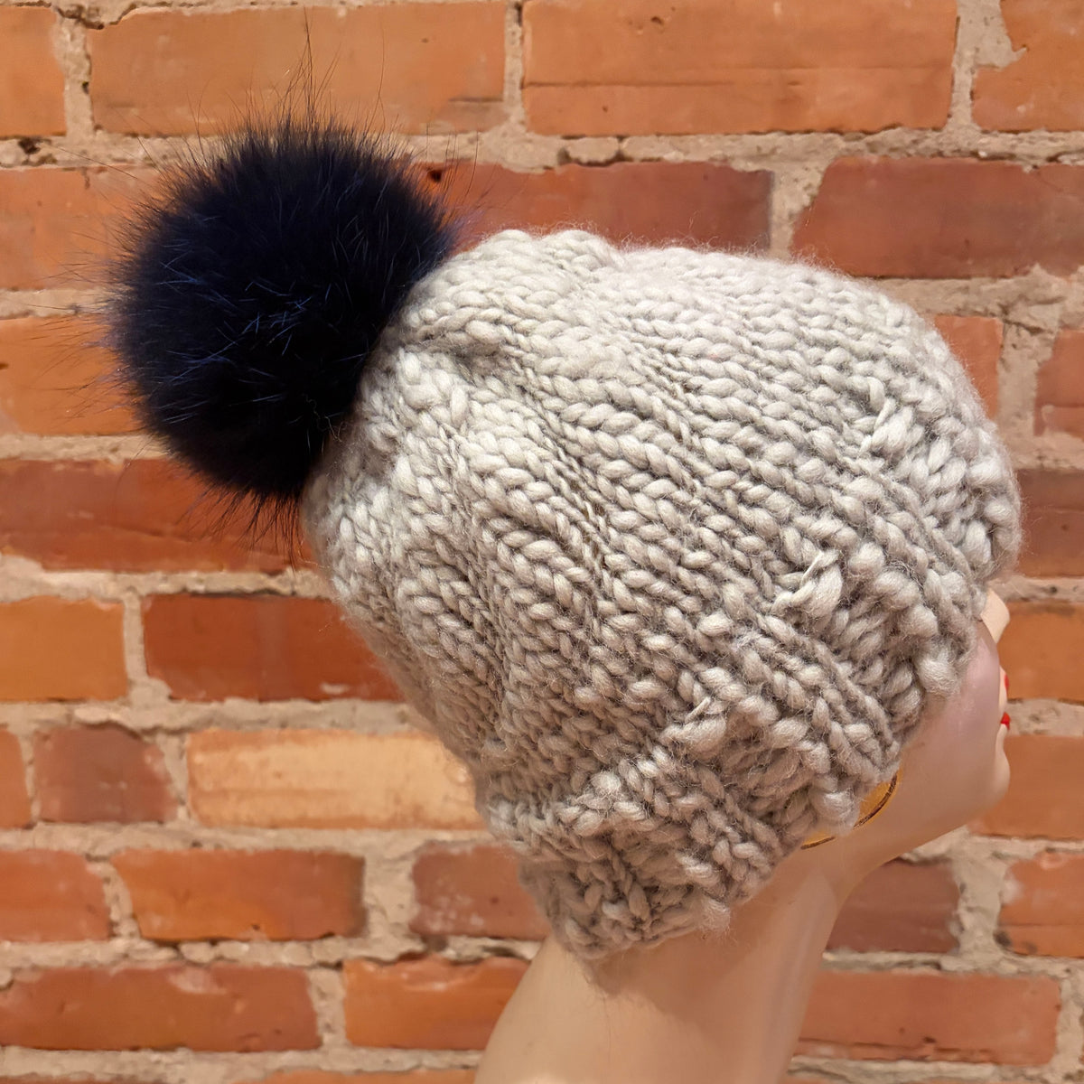 A chunky knit beanie hat in a light oatmeal gray color with a lindigo blue fox fur pom-pom, displayed on a mannequin head against a red brick wall background.