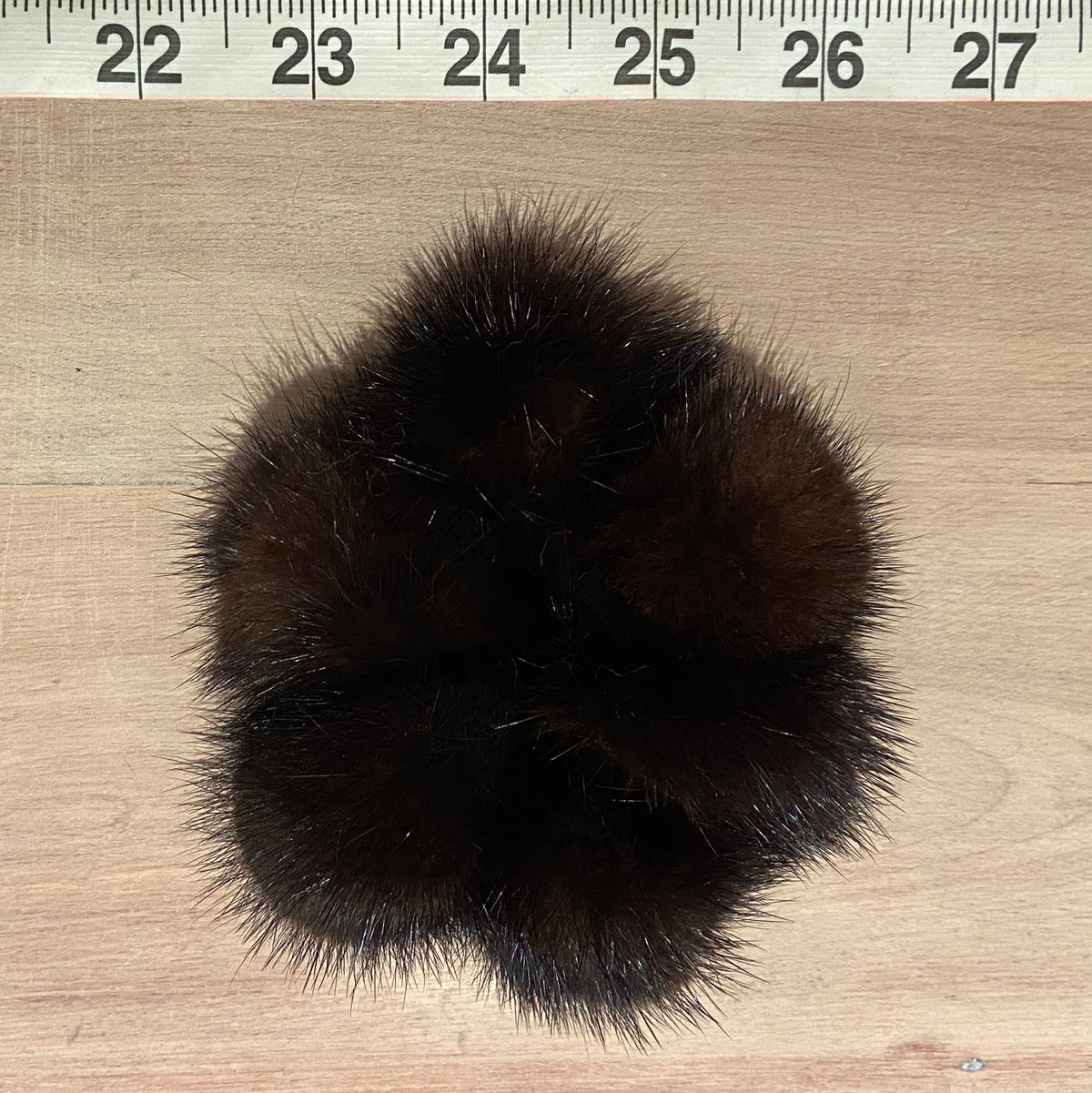 A dark brown fluffy mink fur pom-pom accessory, likely a scrunchie or hair tie, shown on a light wood surface with a measuring tape ruler at the top for scale.