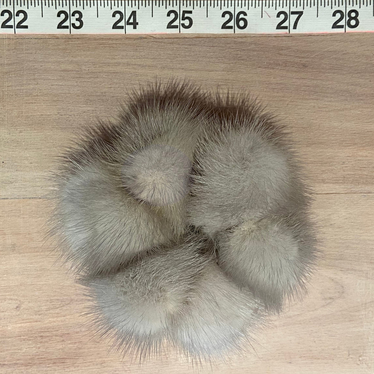 A small fluffy mink-fur scrunchie accessory photographed from above on a light wood surface, with a measuring tape visible along the top edge for scale.