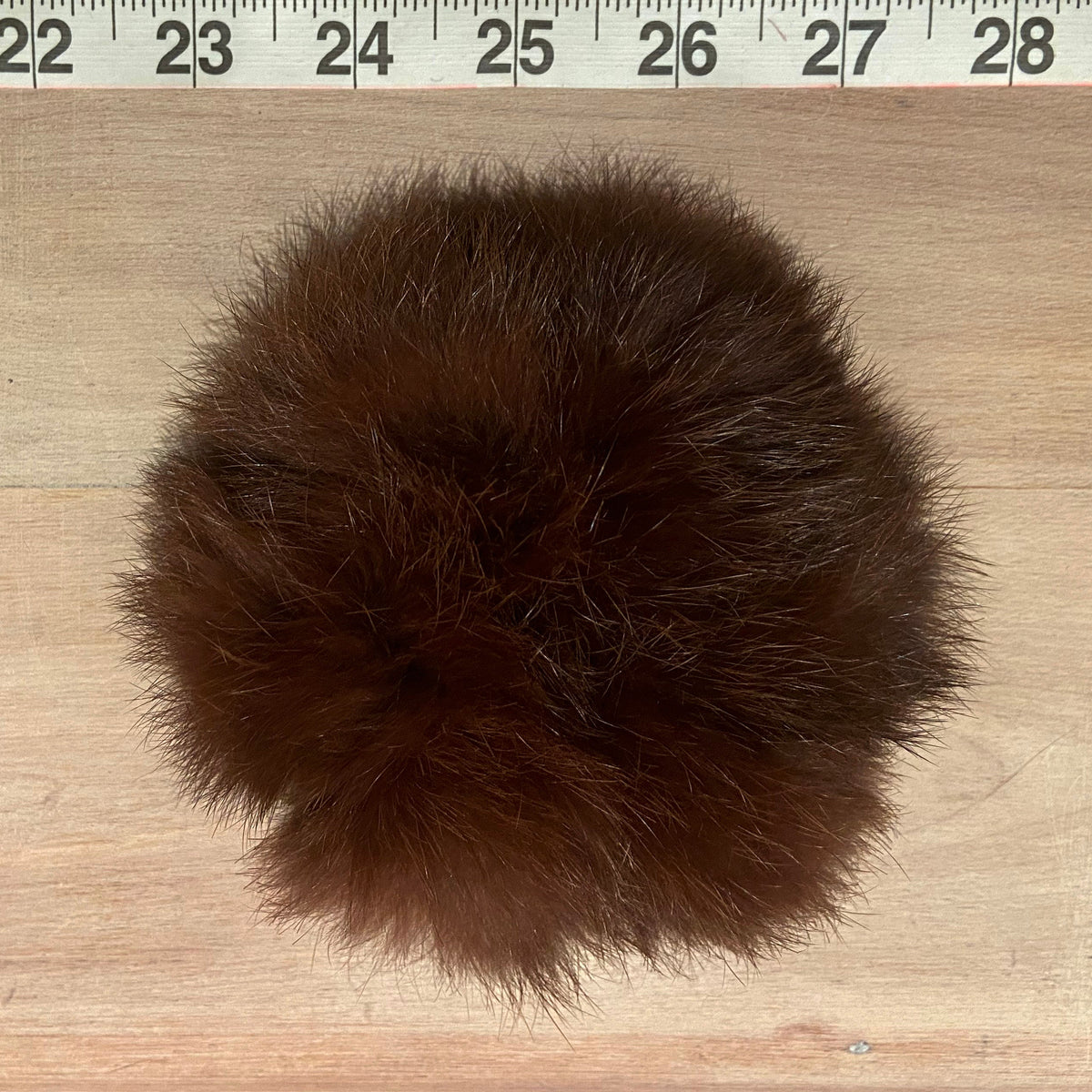 A dark brown fluffy rabbit fur scrunchie placed on a light wood surface with a measuring tape ruler visible at the top edge.