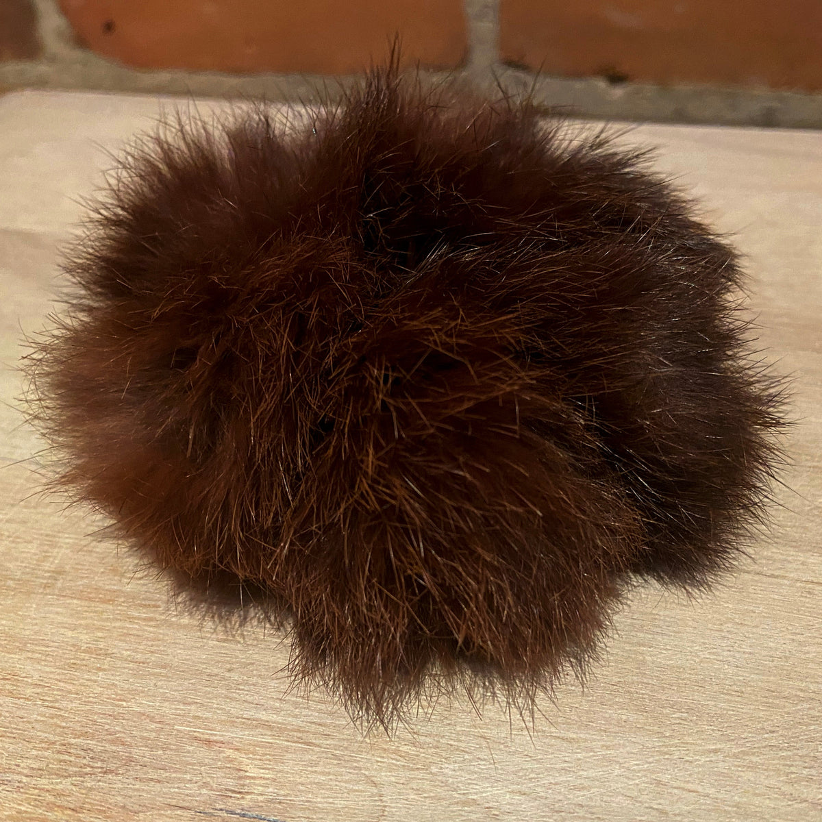 A dark brown fluffy rabbit fur scrunchie accessory resting on a light-colored wooden surface with a brick wall in the background.