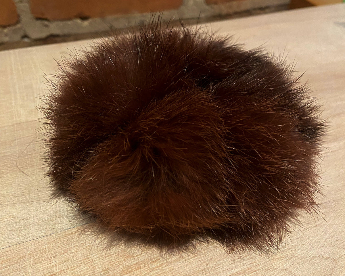 A dark brown fluffy rabbit fur scrunchie accessory resting on a light-colored wooden tabletop with a brick wall in the background.