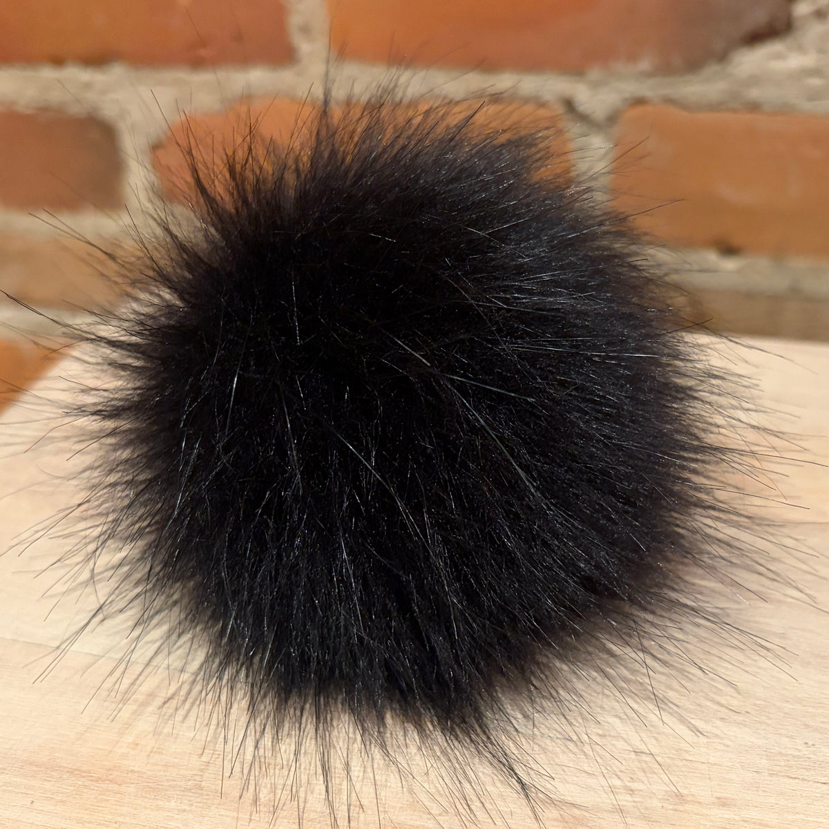 A fluffy black faux fur pom-pom ball resting on a light wood surface with a brick wall background.