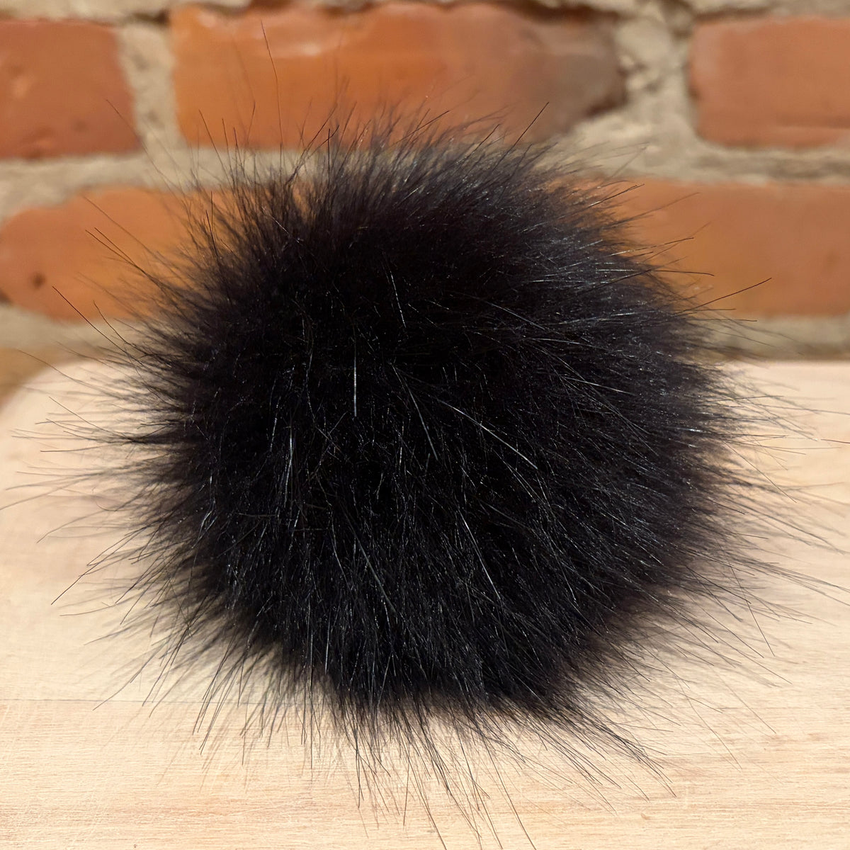 A black fluffy faux fur pom-pom resting on a light wood surface with a blurred brick wall background.